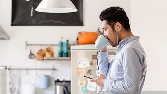 A man with the headphones is standing in kitchen and sipping from the cup 