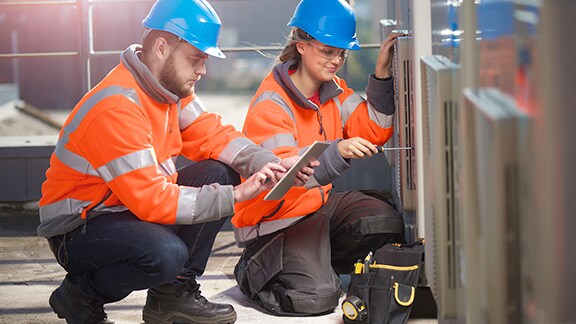 Two maintenance workers kneeling before a device, while one scrolls on a tablet