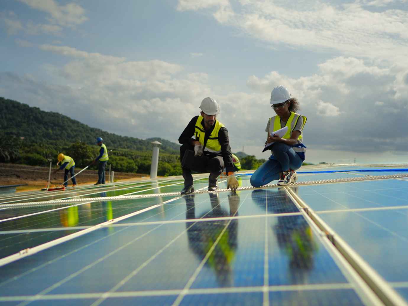 Two engineers examining a solar panel, with more engineers working behind them