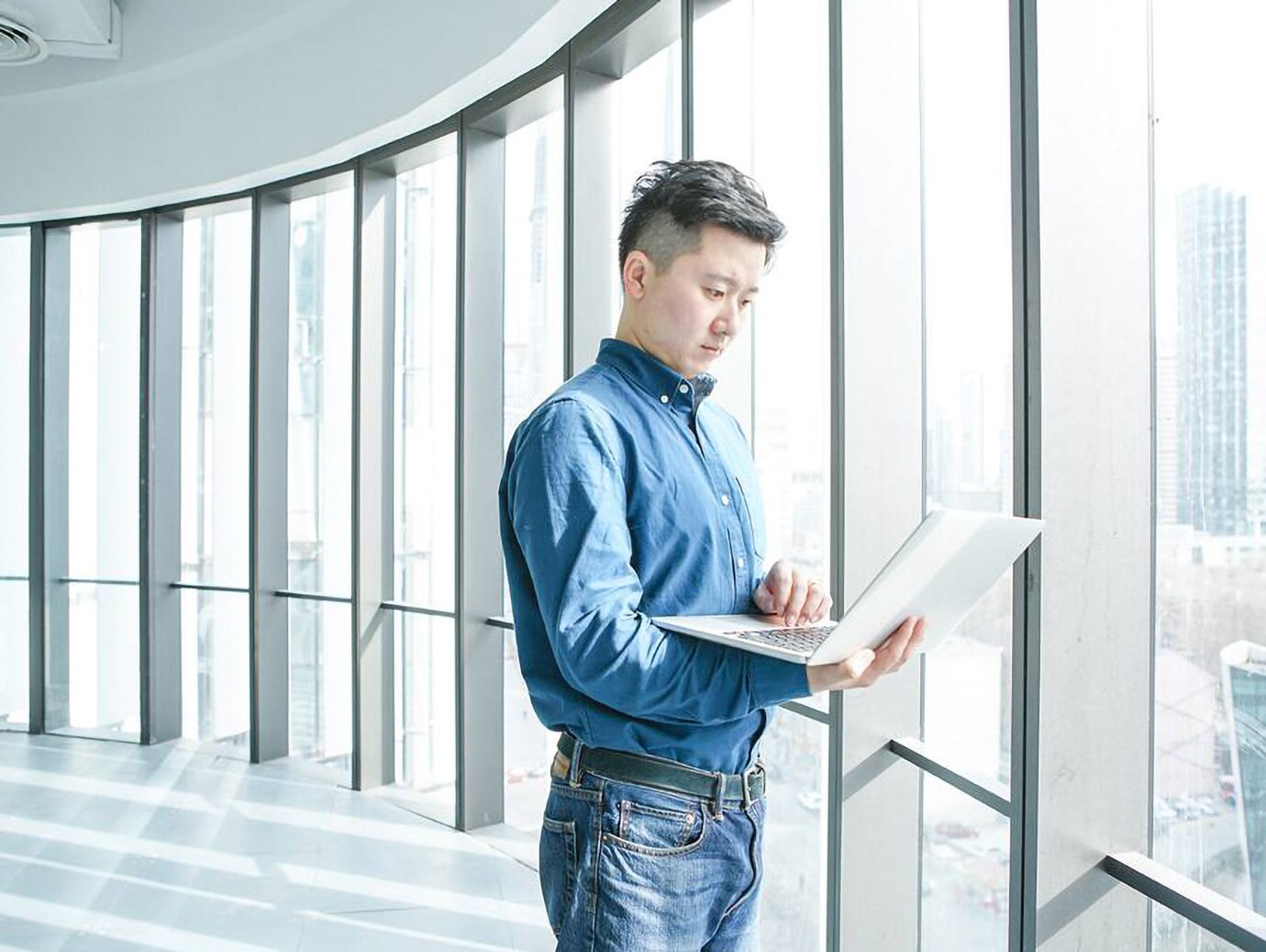 An employee working on a laptop is standing next to a glass window of a corporate office